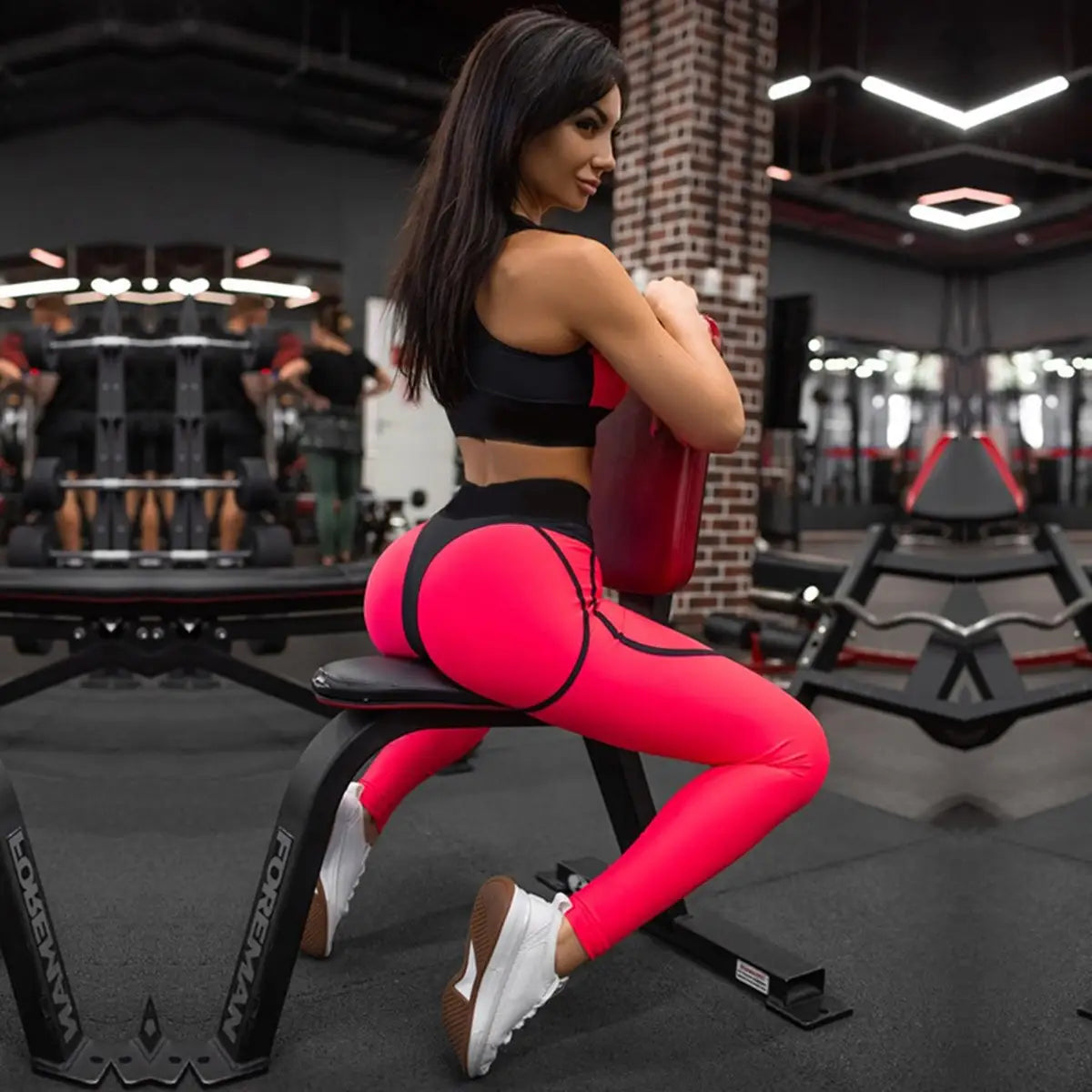 Femme en legging de sport rose vif et soutien-gorge de sport noir, assise sur un équipement d’exercice dans une salle de sport.